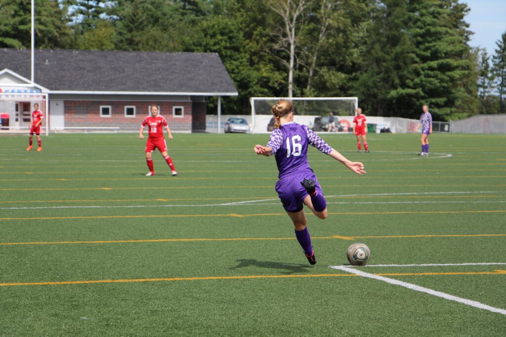 Young woman in Bishop's soccer uniform from the back kicking the ball. Other players in red are further down the field.