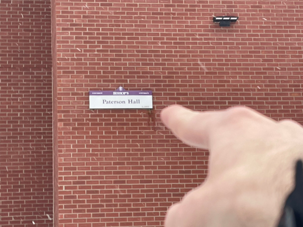 A hand pointing at the sign designating Paterson Hall, on the side of the red-brick residence building.