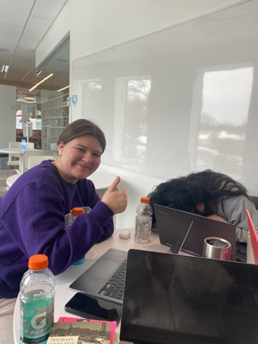 Girl in purple smiling with a thumbs up. Computers and book in front of her.