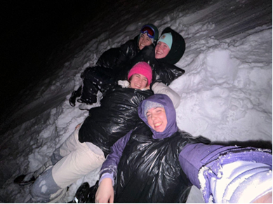 Four girls posing for a selfie while laying down in the snow.