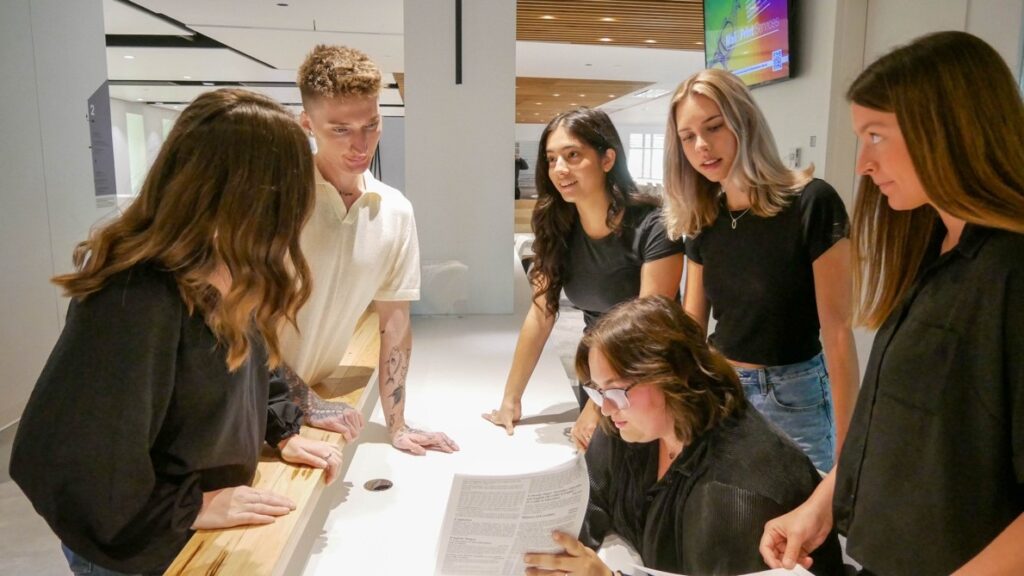 A photo of five people standing around one person sitting at a desk and looking at what seems to be an assignment sheet.